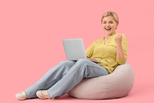 Happy young female programmer with laptop sitting on beanbag chair against pink background