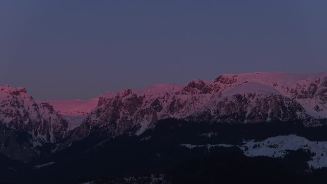 Breathtaking deep pink alpenglow illuminating the rugged snow-capped Bucegi Mountains crest at dusk, tele aerial pan part one, cinematic 4K