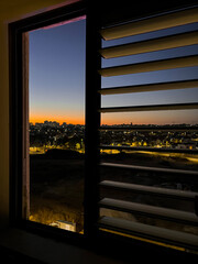 Fototapeta premium Cityscape view through window blinds at twilight, urban horizon with glowing lights and sunset sky, the sunset sky displays a gradient of orange and blue hues