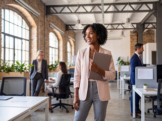 Confident young African American businesswoman holding laptop, smiling while standing in a busy modern office. © Athena Studio