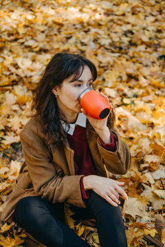 Woman sits on leaf covered ground and drinks from reusable container. Reusable thermal mug, zero waste coffee break, sustainable travel tumbler.