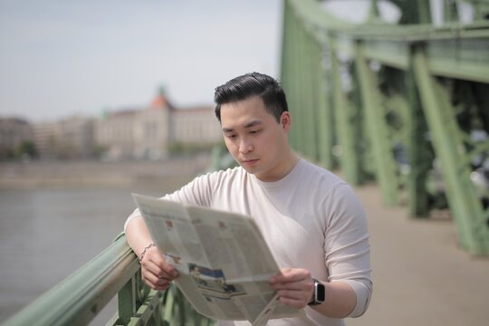 Young Asian man reading newspaper on a city bridge