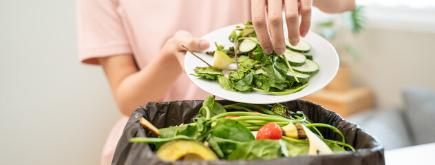 Naklejka premium Person scraping leftover vegetable salad from plate into a trash bin, food waste concept.