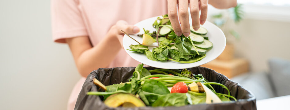 Person scraping leftover vegetable salad from plate into a trash bin, food waste concept.