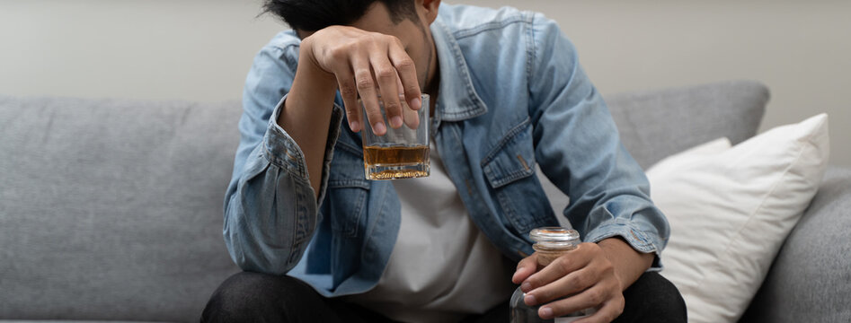 Close-up of depressed young Asian man holding a glass of whiskey, suffering from alcoholism and mental health problems at home.