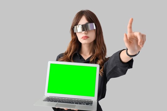 Young woman in futuristic visor holding laptop with green screen and interacting with virtual interface