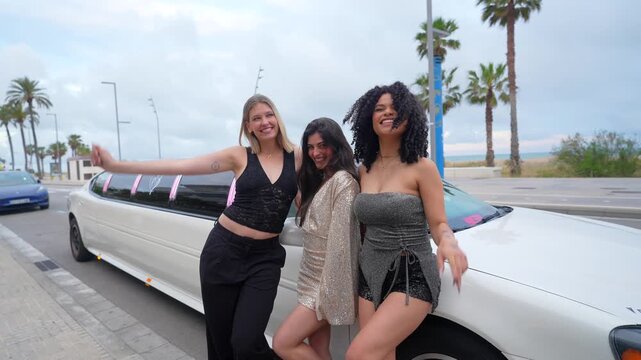 Three happy multiethnic young women in sparkling outfits celebrating a special occasion, laughing and waving at the camera while leaning against a white limousine parked on a street with palm trees