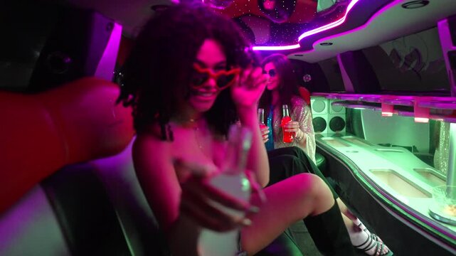 Group of stylish young female friends having fun and drinking inside a luxurious limousine with vibrant neon lights, celebrating a special event like a bachelorette party or prom night