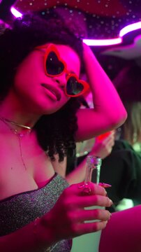 Stylish young woman with afro hairstyle and heart shaped sunglasses dancing and drinking with friends in a limousine, enjoying the vibrant neon lights and luxurious celebration of a party