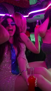 Glamorous multicultural women wearing sparkly dresses and heart shaped sunglasses having fun, drinking, and dancing together inside a limo with vibrant pink neon lights for a special occasion