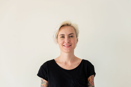 Smiling blonde woman with tattoos standing against a plain white wall