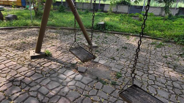 Wide tracking shot of empty vintage chain swings over a stone paved ground in a quiet city park, evoking a nostalgic and lonely afternoon atmosphere.
