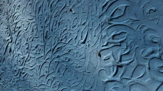 Tracking shot showcasing a rough, light blue painted cement wall with unique relief textures and dynamic tree leaf shadows dancing on the surface.
