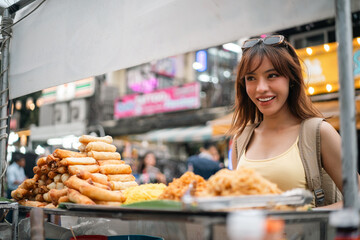 Fototapeta premium Young woman enjoy eating street food at night market. Traveler blogger women Happy tourists Beautiful female with Traditional food local road at thailand bangkok city.