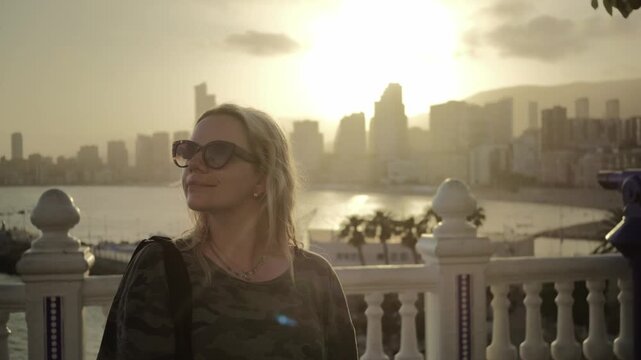 A Blond woman facing the camera and smiling while she is admiring the view in Benidorm, Spain of Mirador del Castell view point in the sunset in slow motion