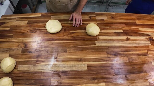 Top-down view of a skilled baker&rsquo;s hands precisely shaping and slapping dough portions on a rustic butcher block, demonstrating artisan breadmaking techniques.