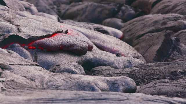 Close-up footage of molten lava from Kīlauea Volcano, Hawaii, slowly advancing beneath a hardened black crust. Bright red magma glows through fissures as the lava cools and reshapes the volcanic terra