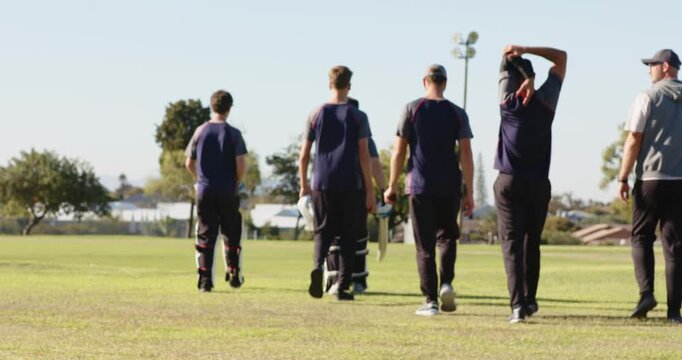 Diverse male cricket teammates walking across outfield carrying bats, gloves after training