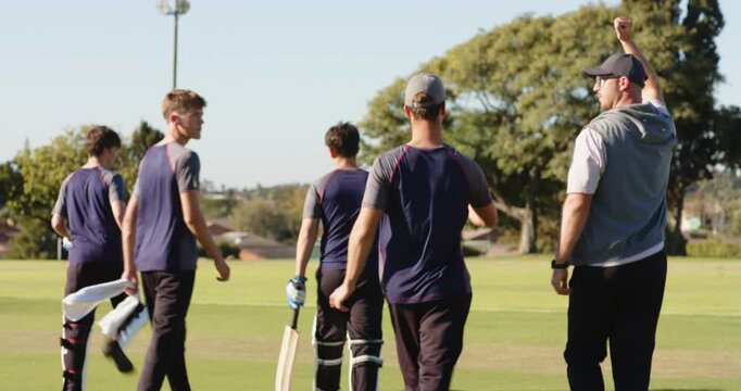 Male cricket team raising arm to signal session end, walking off pitch with bats, pads