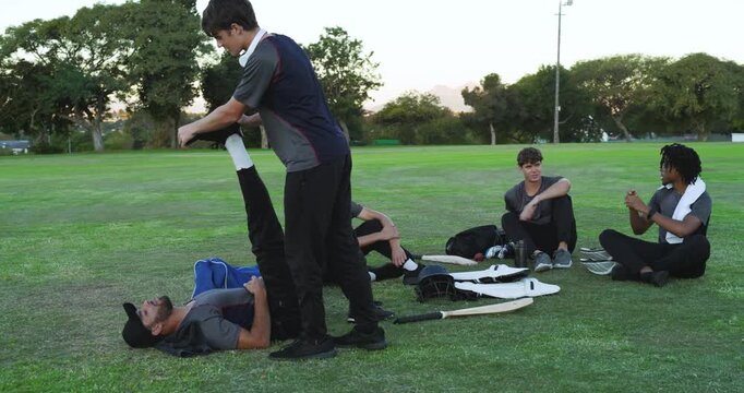 After practice, diverse male teammates stretching, chatting on grass field near cricket bats