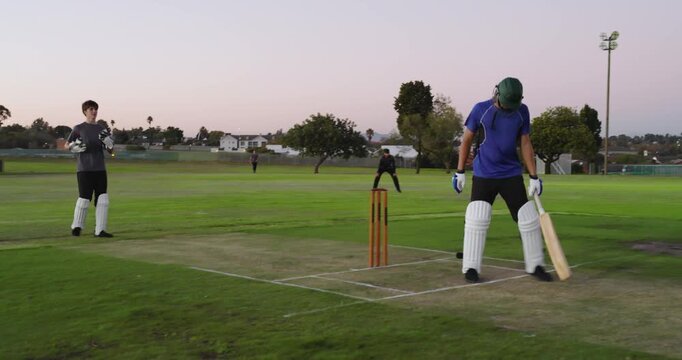 Male batsman hitting offside shot with bat on pitch under floodlight, bowler delivering ball