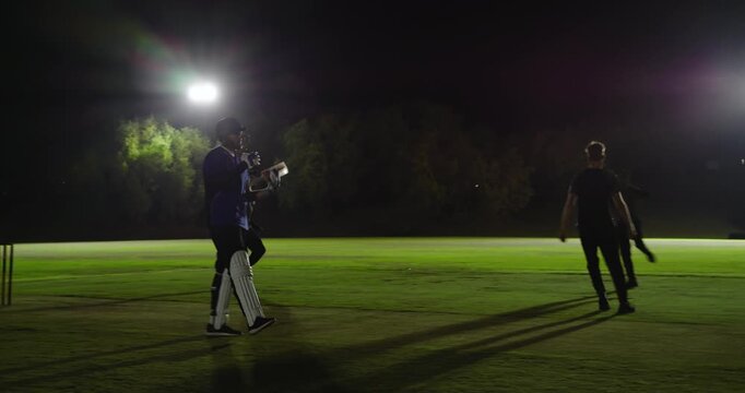 Teenage male bowler bowling ball, floodlights, uprooting stumps dismissing batsman team celebrating