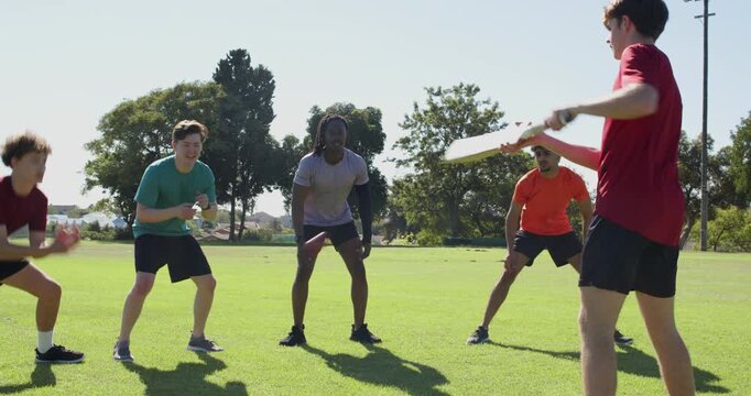Diverse male teammates practicing fielding on field reacting to coach flicking red ball off board