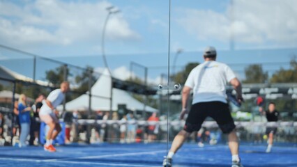 Obraz premium Padel players competing on blue outdoor court viewed through glass wall