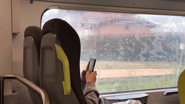 Passenger using smartphone while traveling on train beside rain-streaked window