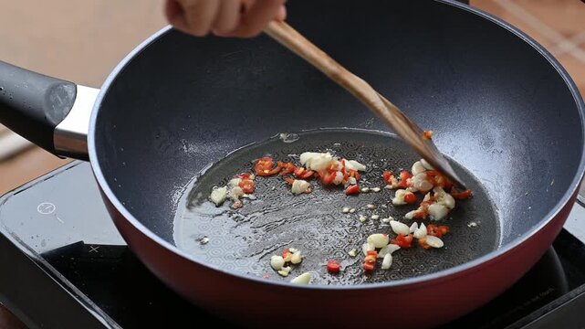 Hand using wooden spatula to stir-fry chopped garlic and fresh chili in hot oil inside a pan, preparing Thai food
