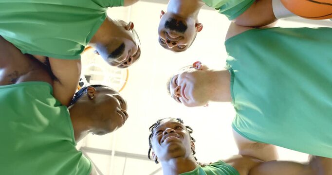 Receiving pep talk, diverse male basketball athletes huddling under hoop in gym breaking out