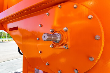 Tight detail of an orange-painted industrial hopper or chute discharge mechanism featuring a central steel shaft, mounting plate, and numerous hex bolts © NastyaPhoto