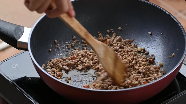 Hand using wooden spatula to stir and mix minced pork with soy sauce and seasoning inside a hot pan
