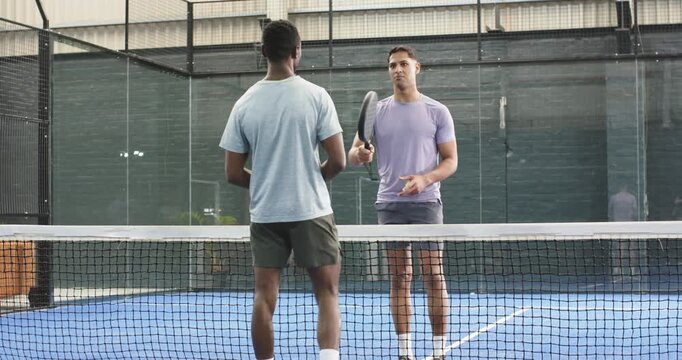 Diverse male friends tapping paddles at net, tossing yellow ball, exchanging volleys on court