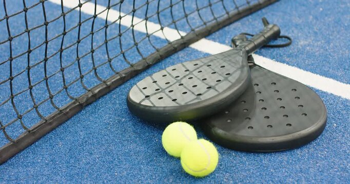 Camera capturing two black padel rackets and balls resting on blue turf under taut net
