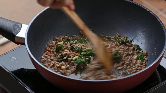 Hand using wooden spatula to stir and mix holy basil leaves and minced pork inside a hot pan, cooking Thai Pad Ka Prao