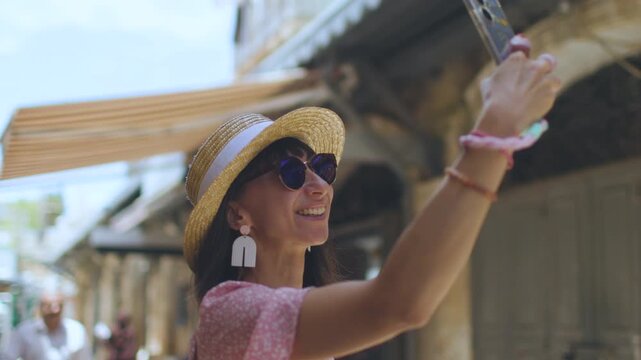 A cheerful woman wearing a straw sun hat and sunglasses holds a smartphone up for a selfie on a bright urban street. People walk in the background in an arched building.