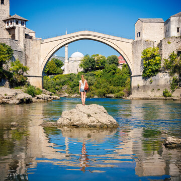 Young female tourist admiring the historic Stari Most bridge in Mostar, beautiful reflection in Neretva river, authentic travel experience in Bosnia and Herzegovina