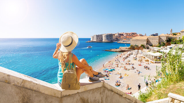 Young female tourist in straw hat looking at the old town of Dubrovnik and Banje beach, authentic summer vacation and travel experience in Croatia
