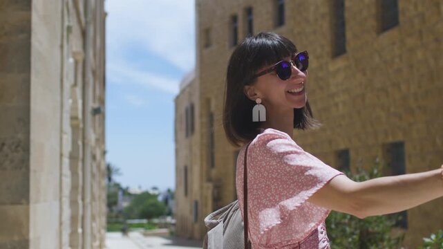 A cheerful woman stands on a narrow urban street and holds a smartphone up for a selfie. She wears reflective sunglasses, a pink patterned dress, and dangling earrings. 