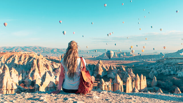 Young female backpacker sitting on cliff watching hot air balloons flying over Cappadocia, authentic dream travel experience and wanderlust concept in Turkey