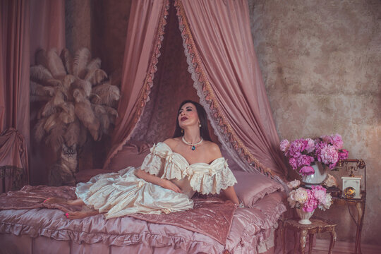 Elegant mature woman in her 40s or 50s in a luxury vintage cream dress relaxing on a pink bed with a canopy, beautiful middle aged lady in a romantic boudoir with peony flowers