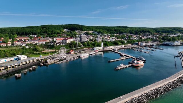 Aerial footage of Sassnitz harbor on the island of R&uuml;gen, Germany with marina facilities, passenger vessels, tourist ferris wheel and waterfront town backed by forested hills.