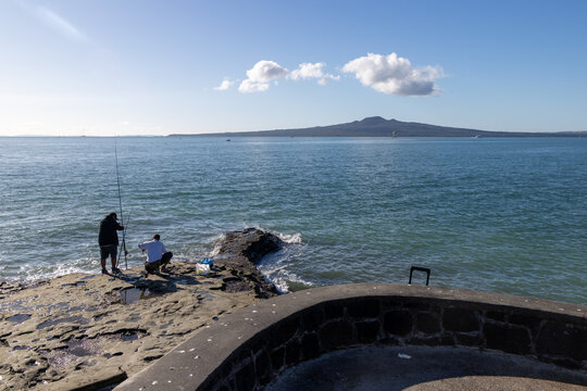 Two men fishing from a rocky shore in Mission Bay Auckland, New Zealand, with Rangitoto Island in the background on a clear day.