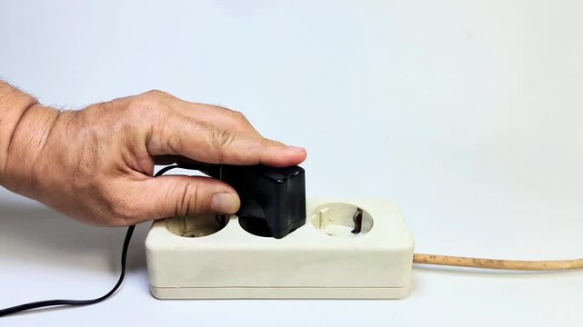 A man's hand tries to plug a black charger adapter into an electrical socket on a white background.