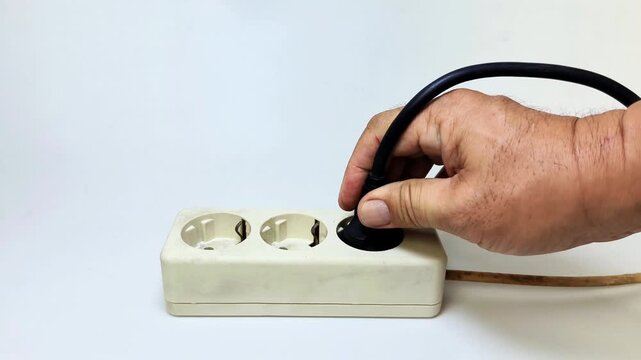 A man's hand tries to plug a black power cord into an electrical socket on white background