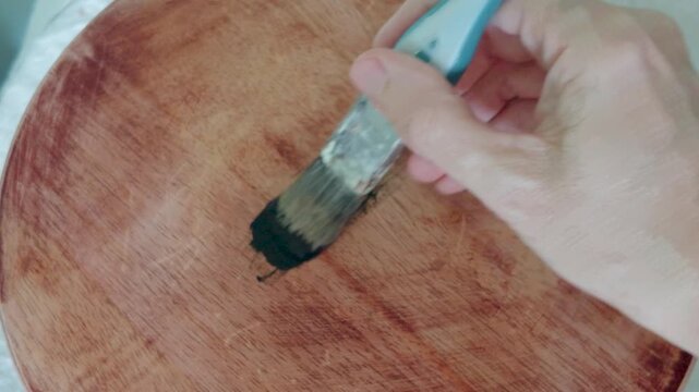 Painting sanded table with black paint as a brush coats raw wood surface in a DIY furniture upcycling and home improvement project, close-up view of craftsmanship and renovation work.