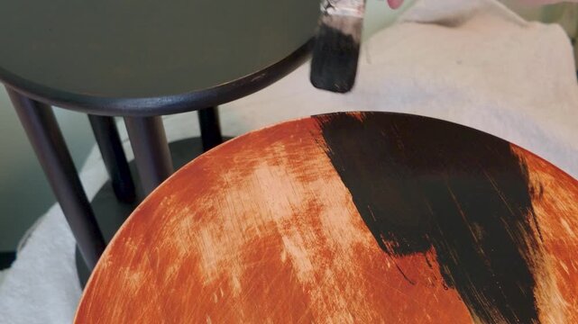 Painting a sanded wooden side table with black paint using brush strokes, DIY furniture makeover indoors; a matching table sits behind already coated and drying.