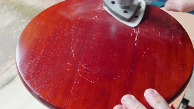 Multitool sander smooths cherry red round table top in close-up. DIY furniture refinishing, surface prep, removing varnish and scratches for repaint or restain.