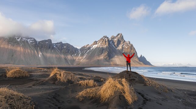 Man in red jacket stretches arms into the air, black lava beach, sandy beach, dunes with dry grass, mountains Klifatindur, Eystrahorn and Kambhorn, headland Stokksnes, mountain range Klifatindur, Austurland, East Iceland, Iceland